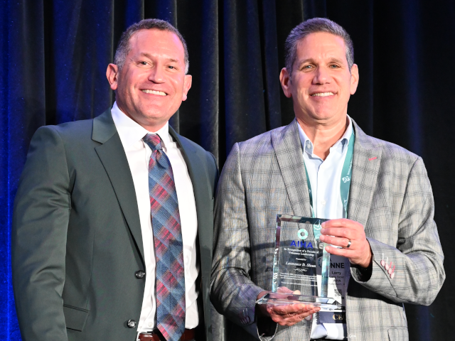 A photo of two white men in suit jackets standing in front of a blue curtain. Larry Sloan, at right, has just received a clear glass award from Bradley King, at left.